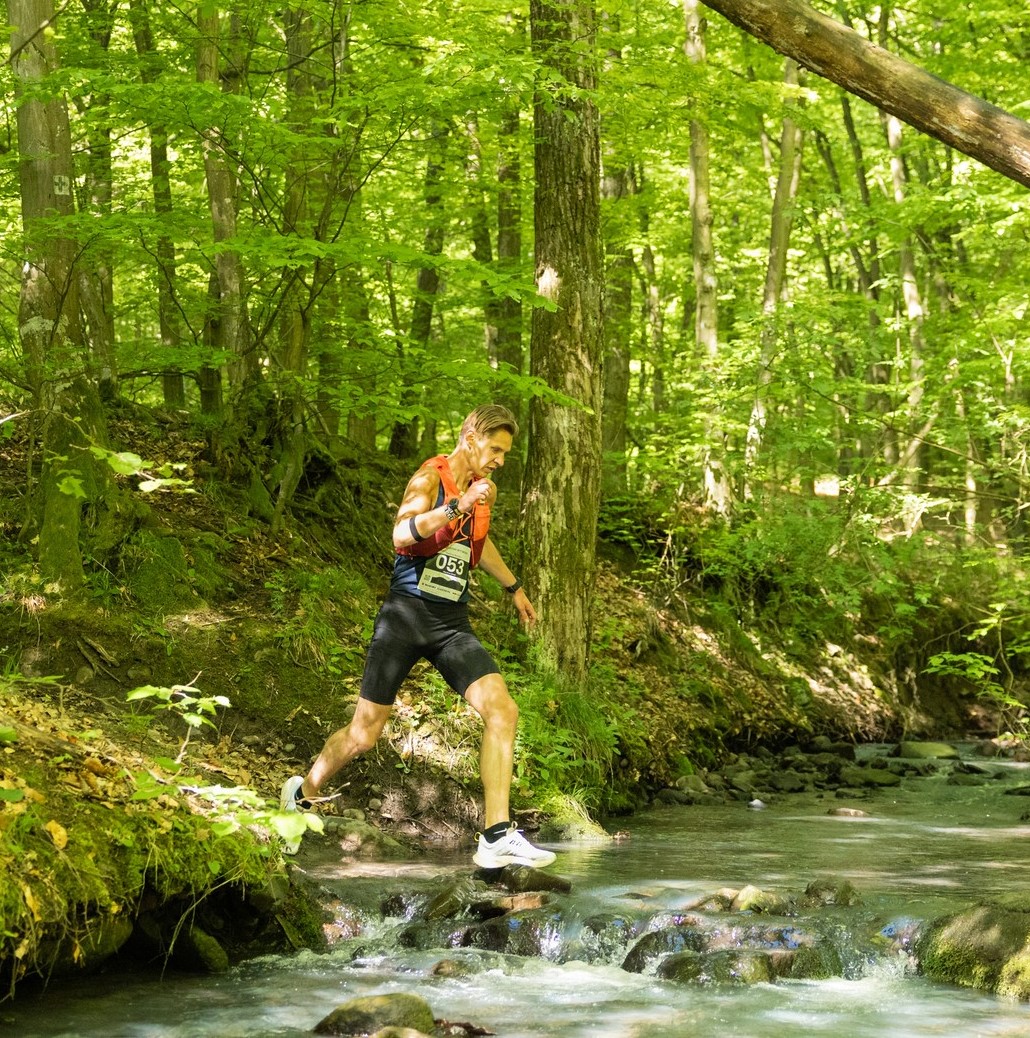 Berci crossing a rocky stream on a forest trail race wearing a race bib